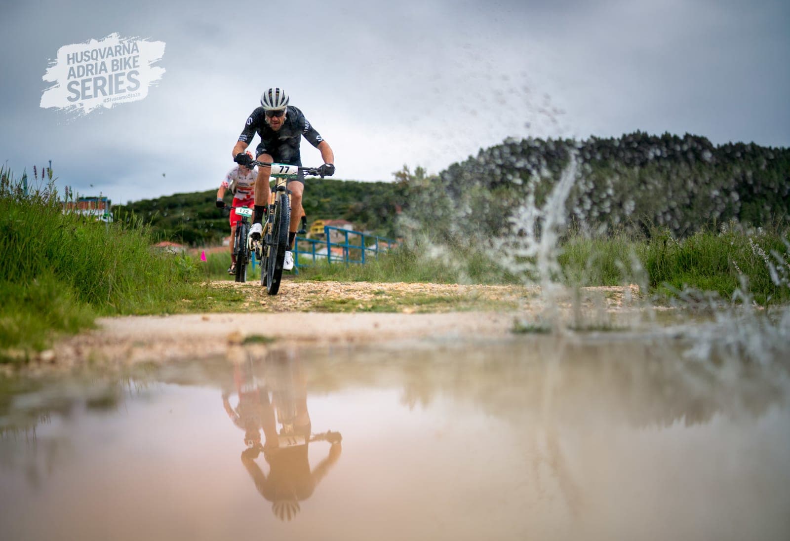 Filip riding through a wet Rab MTB Marathon section with a large puddle reflection in the foreground.