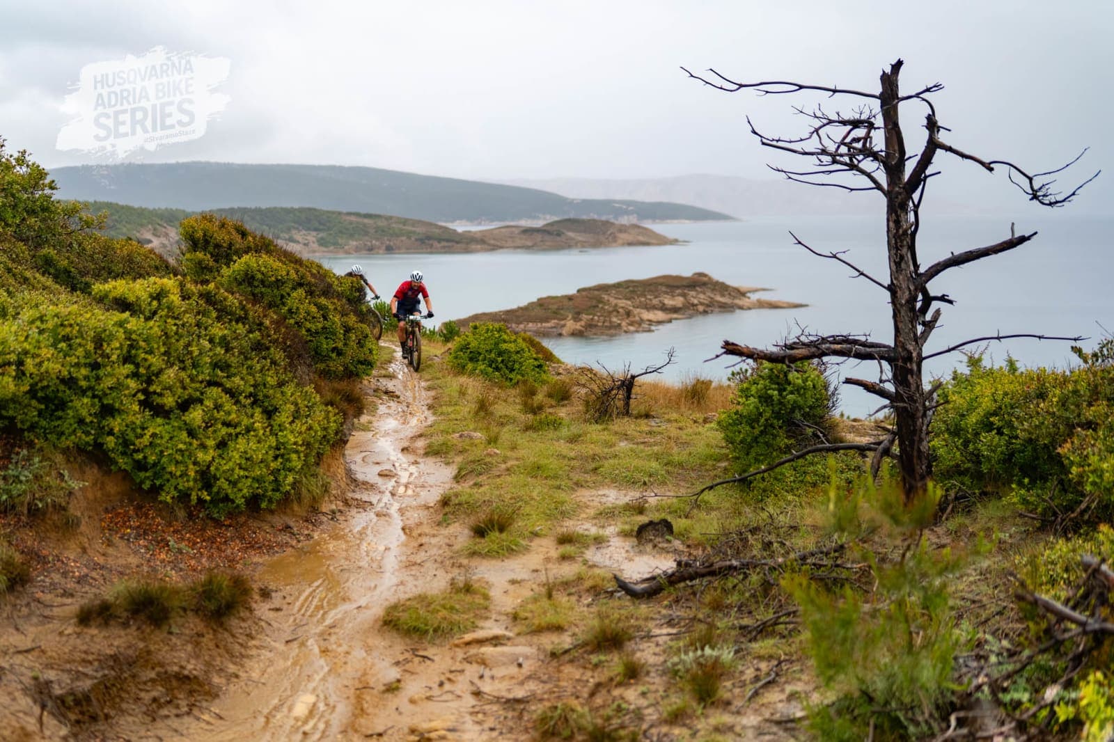 A rider pushing through a muddy coastal singletrack section with the sea behind.