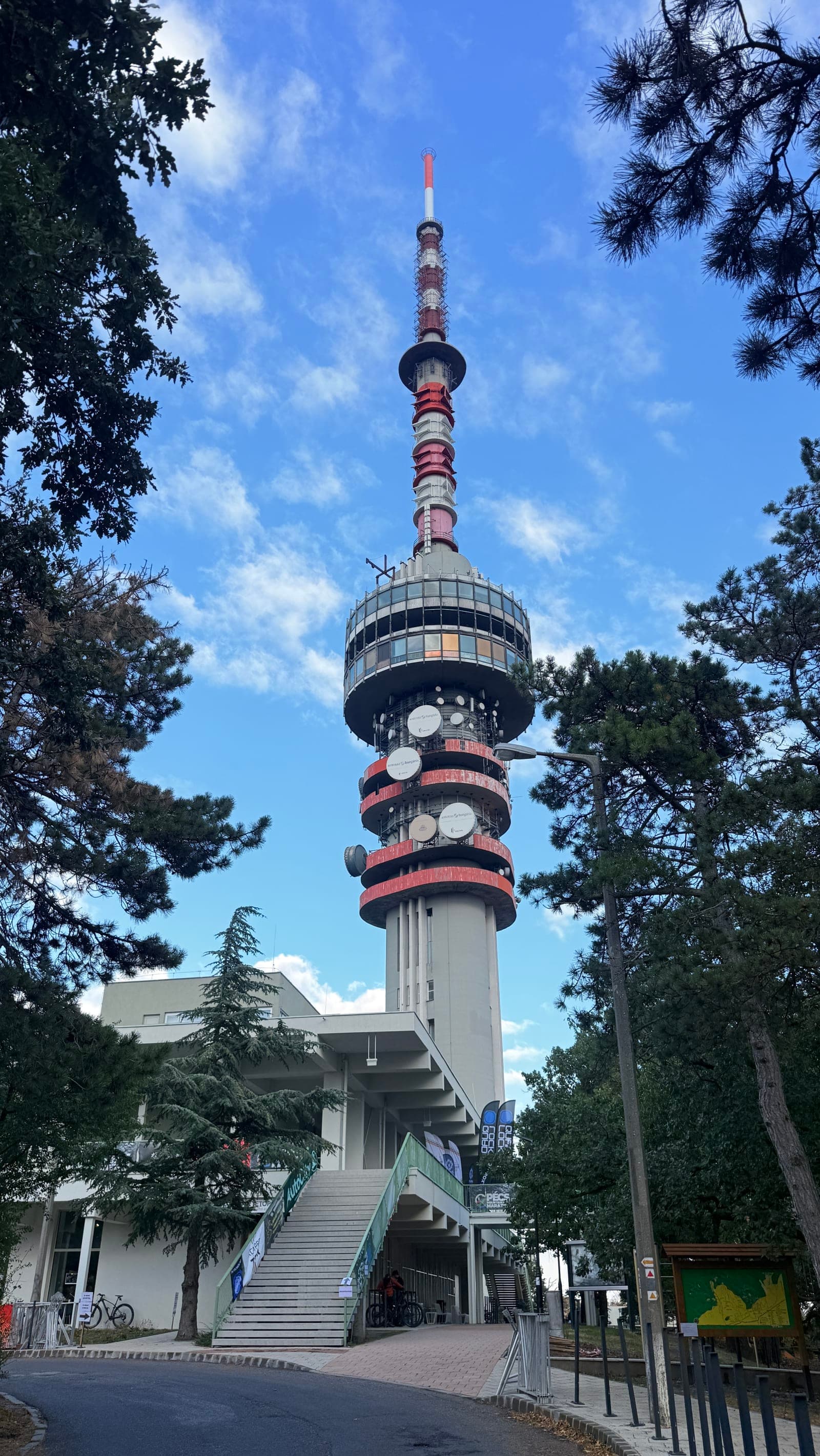 The red and white Pécs TV Tower rising above pine trees, with race banners at the base at the Pécs MTB Marathon.