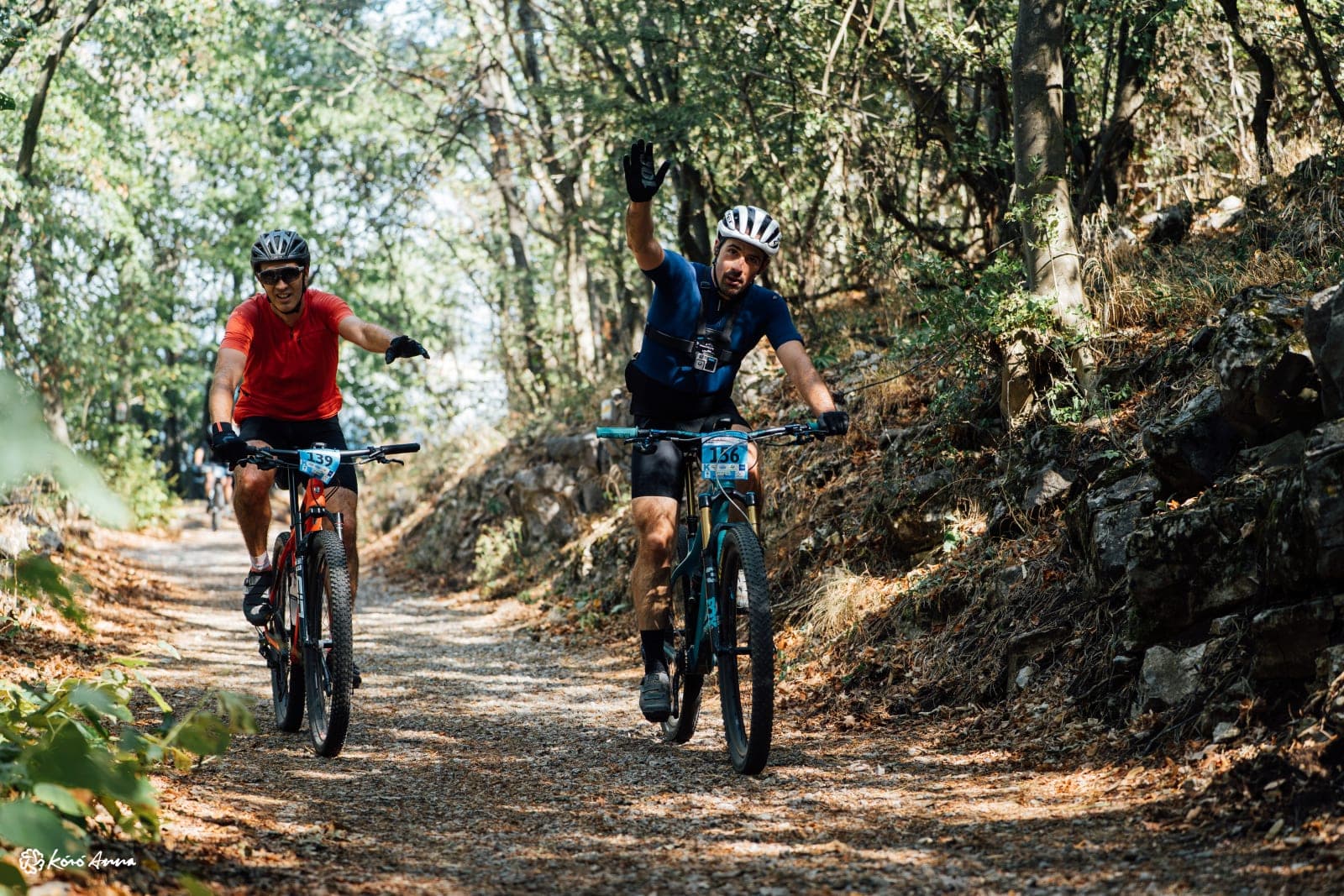 Two mountain bikers riding a gravel forest track in the Mecsek, one in a blue jersey raising a hand toward the camera, during the Pécs MTB Marathon 2026.