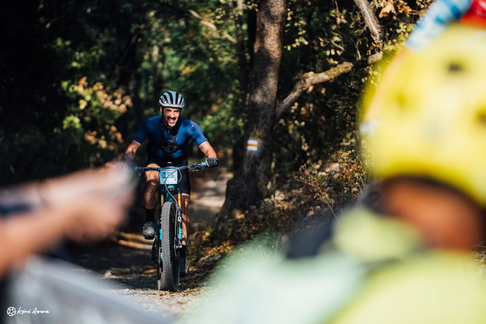 A smiling mountain biker in a blue jersey riding toward the camera on a wooded trail at the Pécs MTB Marathon 2026, with blurred spectators in the foreground.